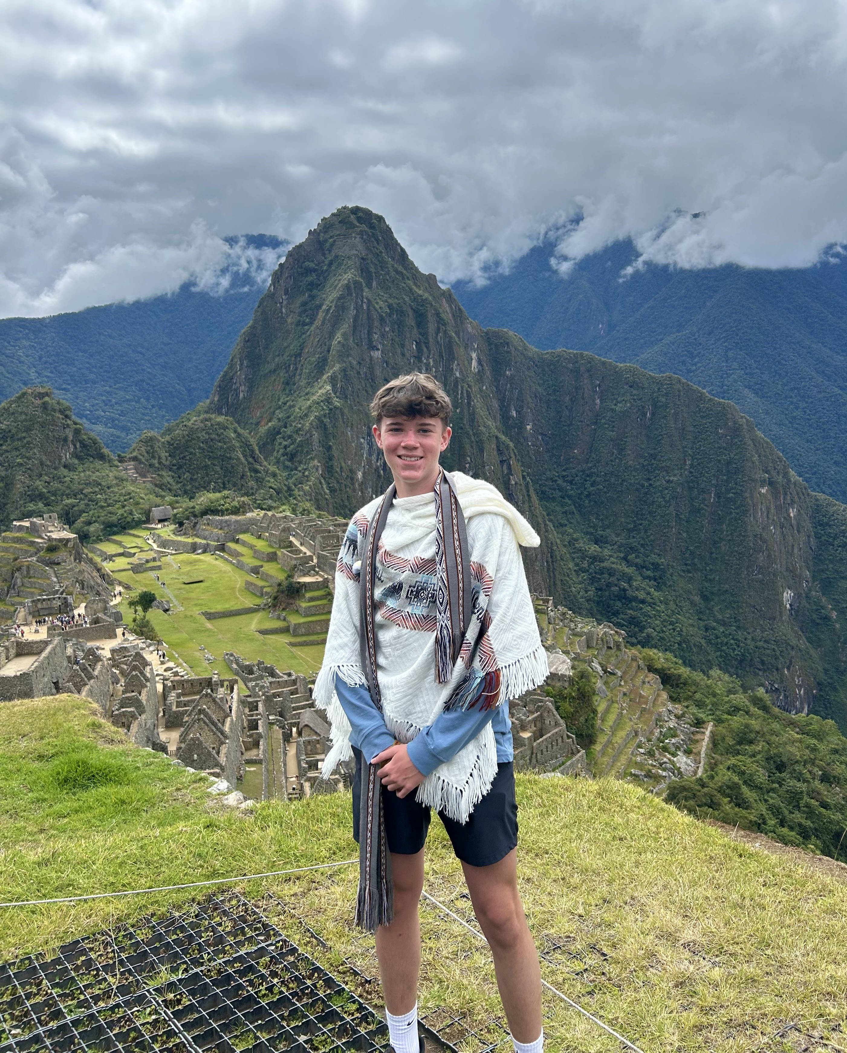Noah Cowley at Machu Picchu, wearing a woven poncho, with Huayna Picchu behind